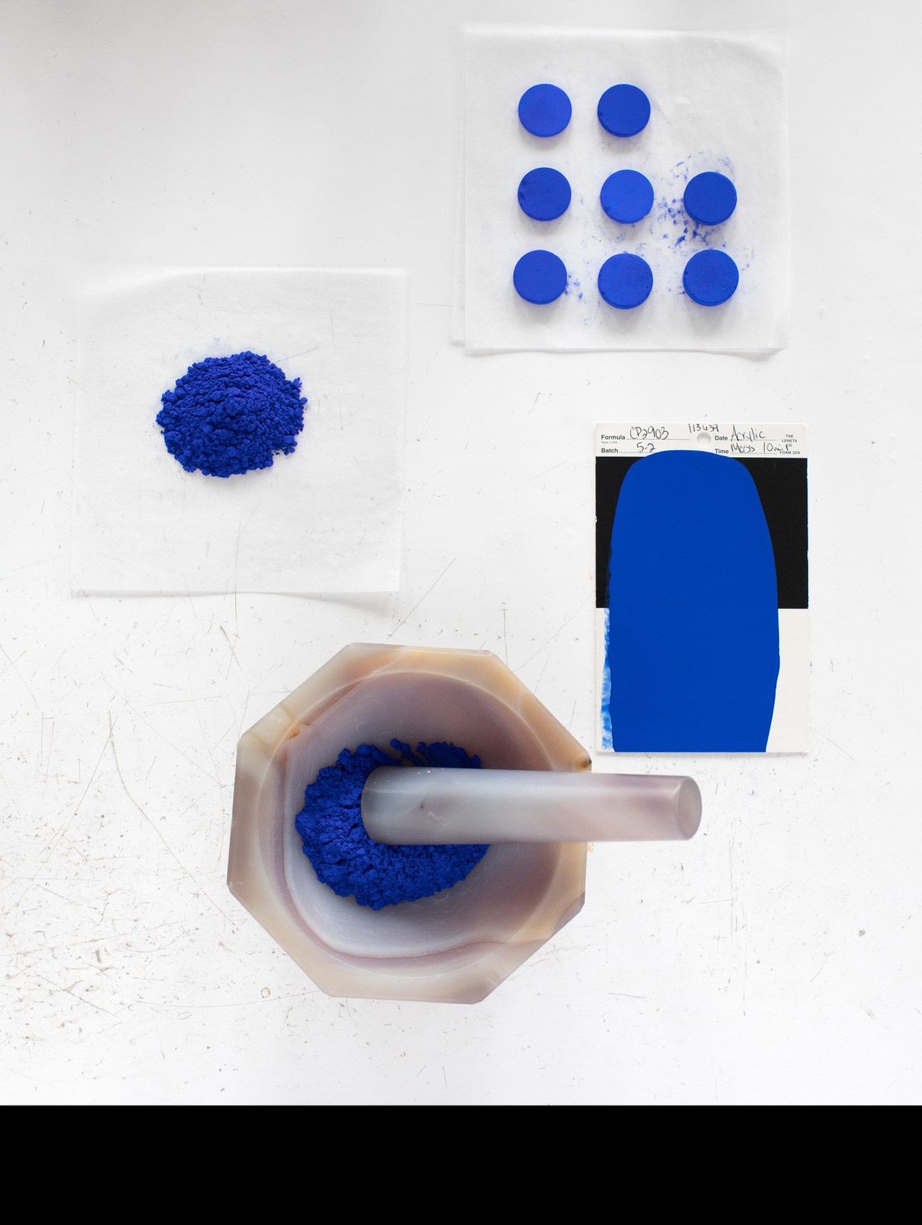 Blue pigment powder in a mortar being ground with a white pestle, with blue pellets and color swatches visible above.
