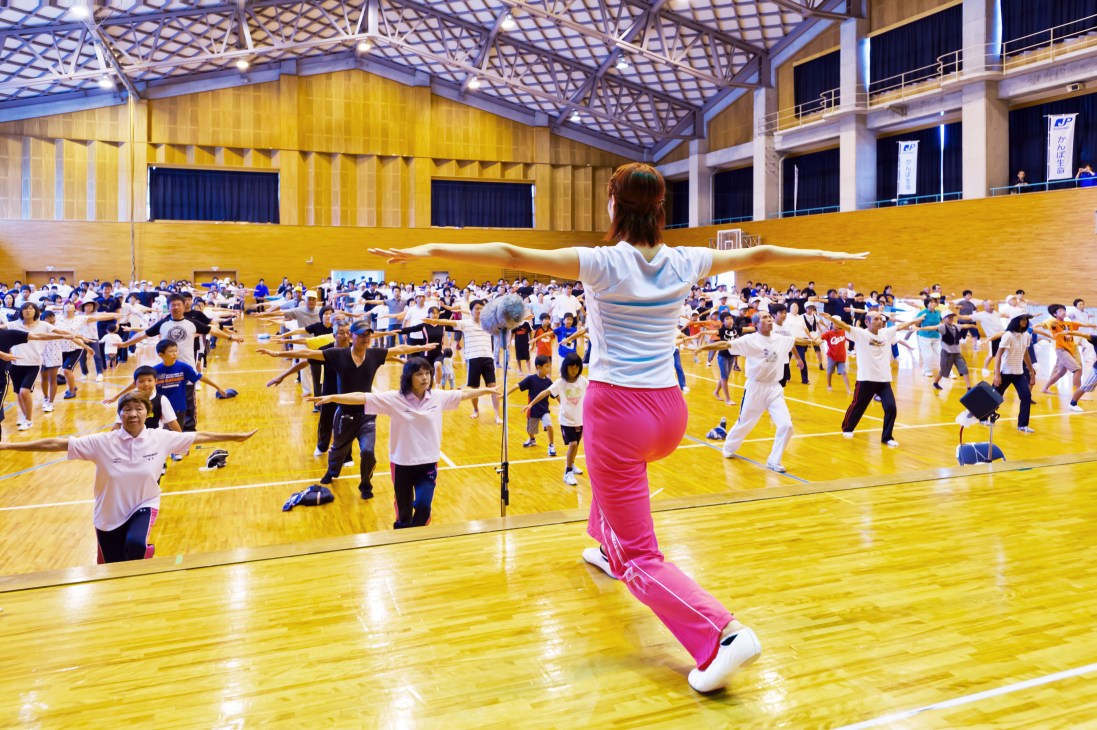 An instructor in pink sweatpants leads 800 people doing rajio taiso exercises in a gymnasium with yellow wooden flooring.