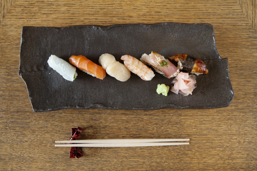 Six pieces of nigiri sushi arranged on a dark slate plate with wasabi and pickled ginger, served with chopsticks.