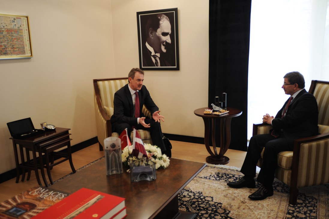 Two men in suits discuss at a formal meeting table with Turkish and Latvian flags, beneath a portrait of Atatürk.