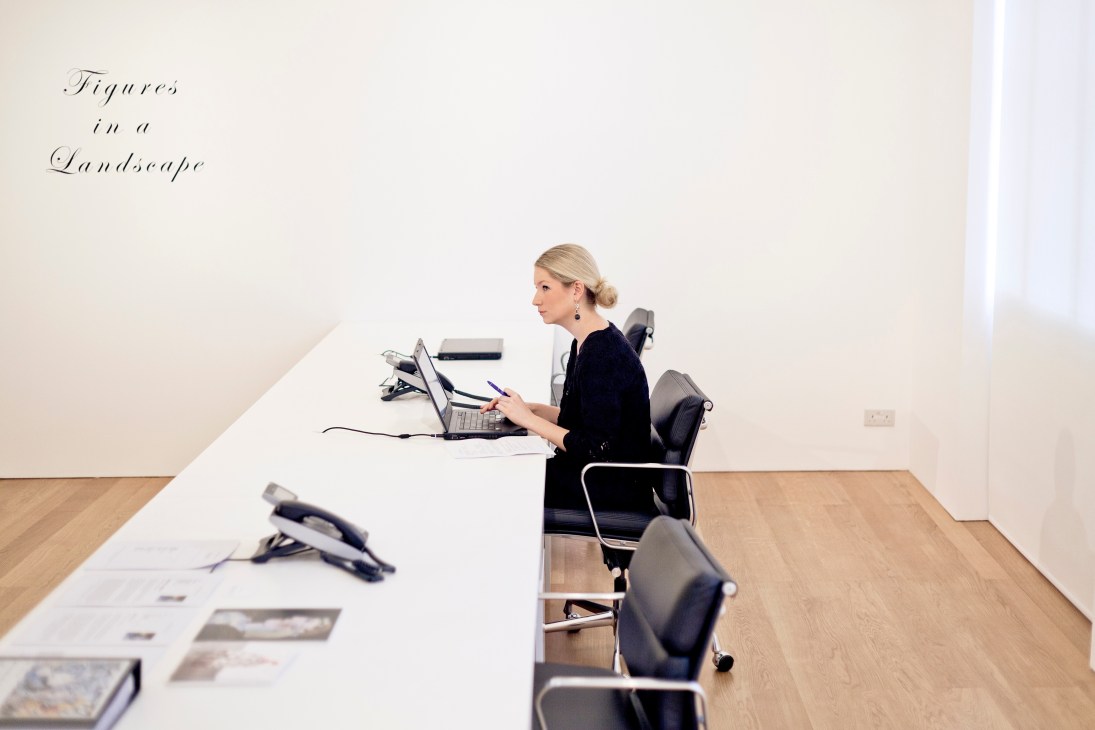 Woman in black blazer sits at white desk with computer in minimalist gallery space.
