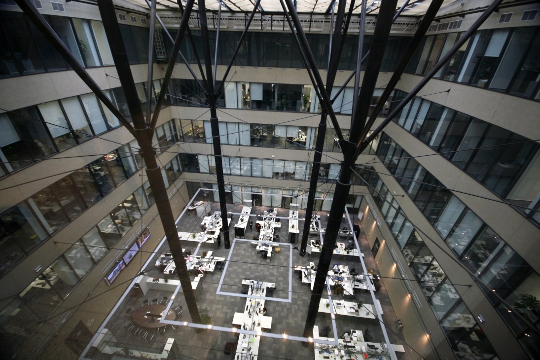Interior view of a multi-story atrium with large black structural beams, white workstations, and glass office windows surrounding the open space.