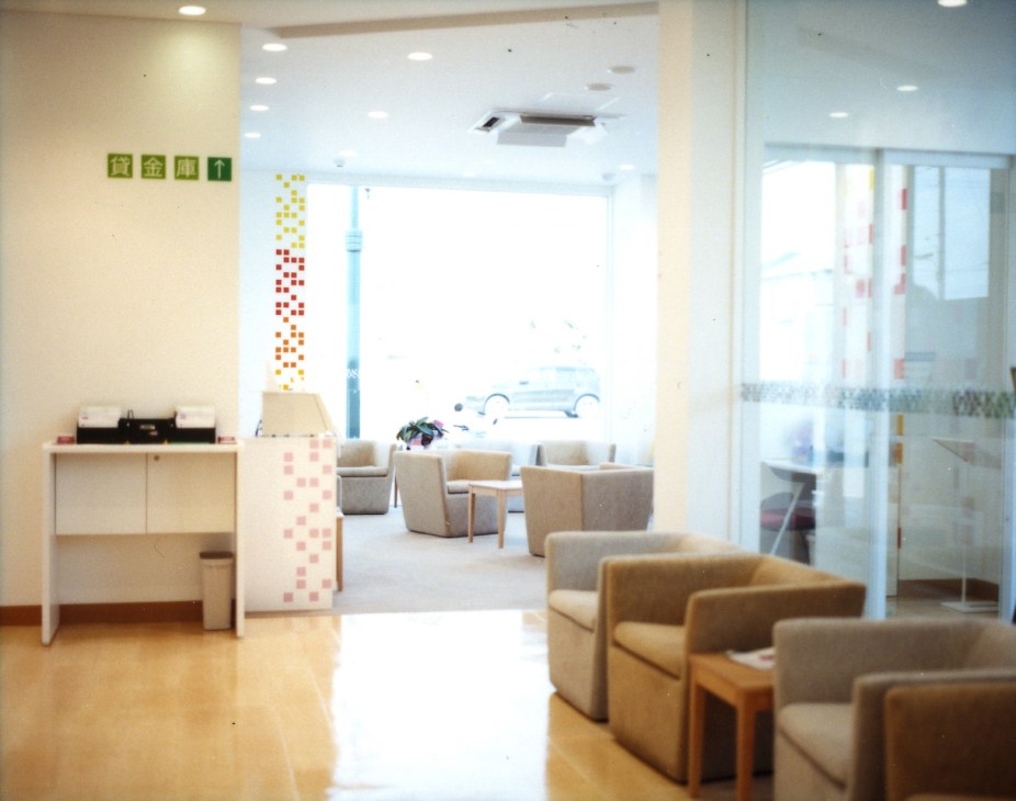Bright interior seating area of Sugamo Shinkin Bank's Niiza branch with beige armchairs, colorful square signage on wall, and large windows.