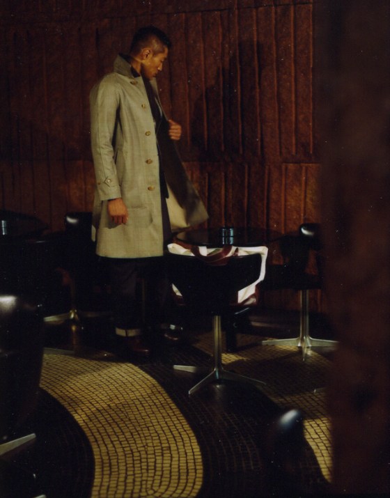 Man wearing a light Mackintosh coat and dark jeans stands in a vintage-style interior with wooden paneling.