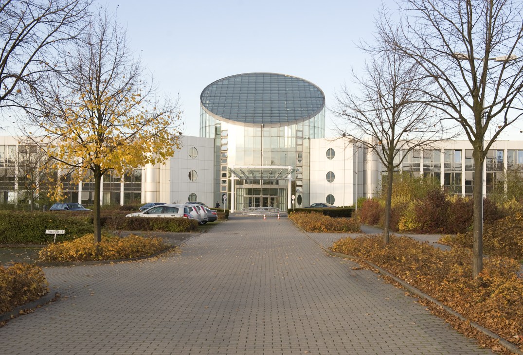 Modern white building with curved glass dome roof surrounded by bare trees and parked cars in autumn.