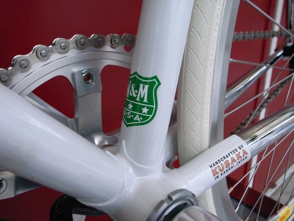 Close-up of white Kinfolk bicycle frame showing chainring, headbadge and handcrafted details against red background.
