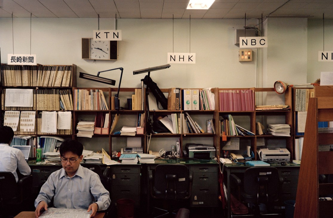 A man works at a desk in Nagasaki city hall's press room, surrounded by shelves of media archives and broadcast clocks.