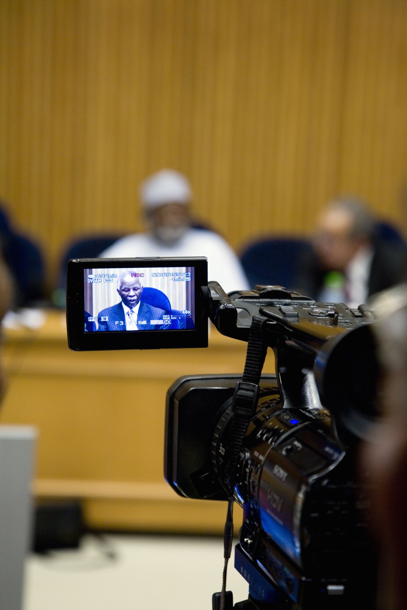 A video camera's monitor displays a speaker at an African Union proceedings, with blurred delegates in the background against a yellow wall.