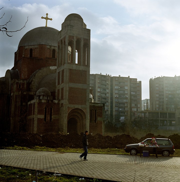 Unfinished Orthodox church with golden cross and bell tower under construction, surrounded by concrete apartment blocks in Pristina.