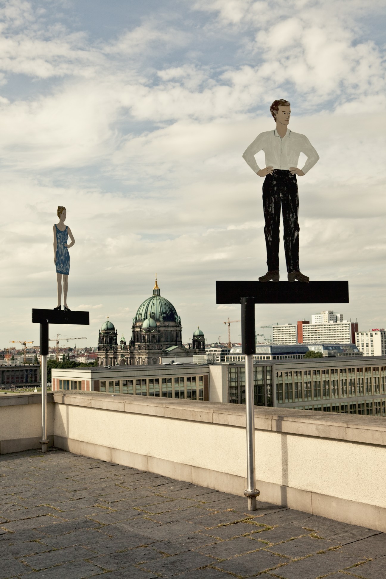 Two illustrated figures stand on pedestals on a Berlin rooftop overlooking the Berliner Dom cathedral.