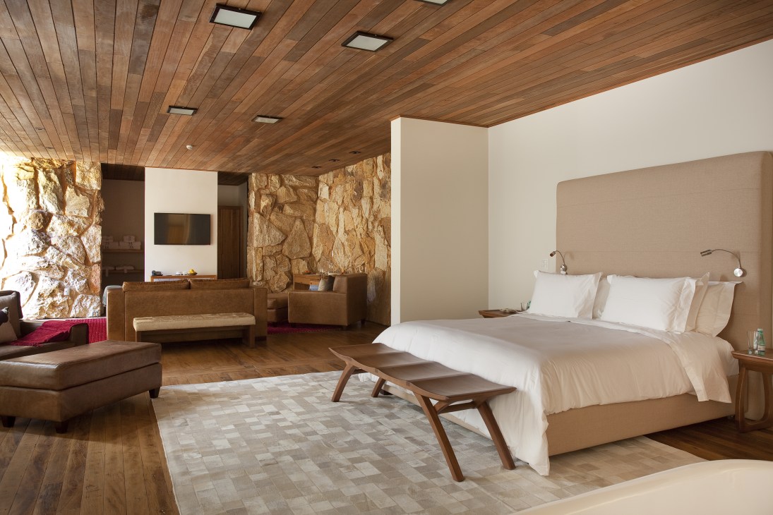 Botanique Hotel bedroom with wooden ceiling, stone walls, bed with white linens, and mid-century modern furniture.