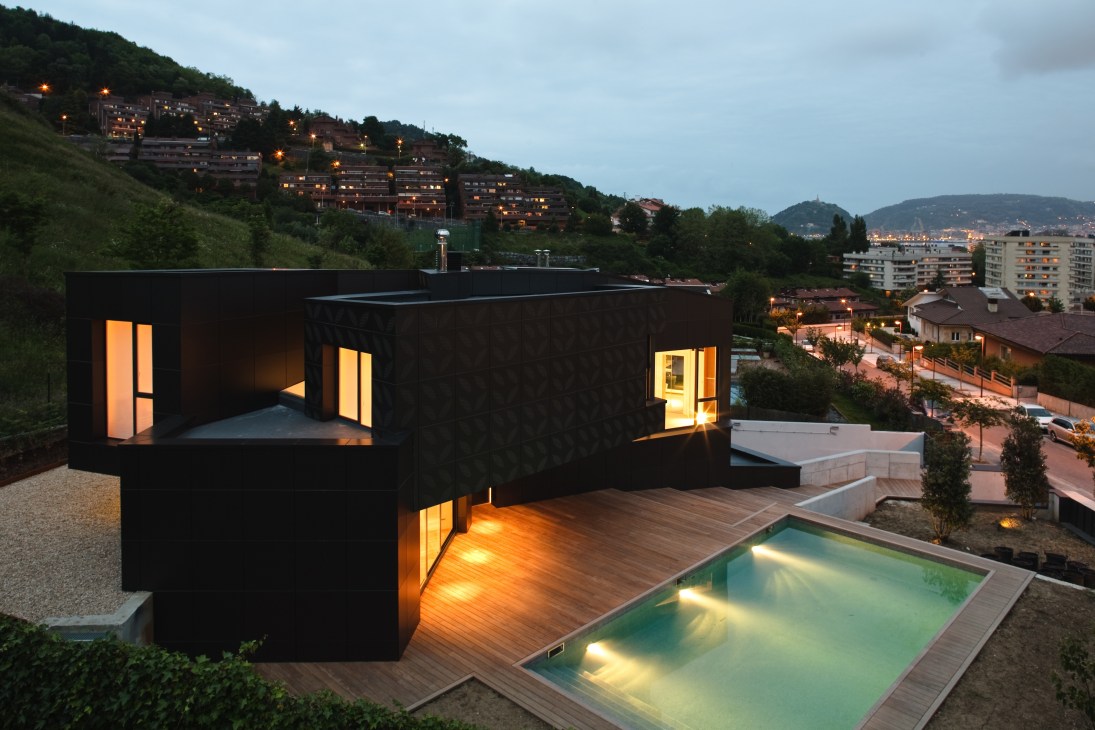 Modern black residence with illuminated windows, wooden deck, and pool overlooking a hillside town at dusk.