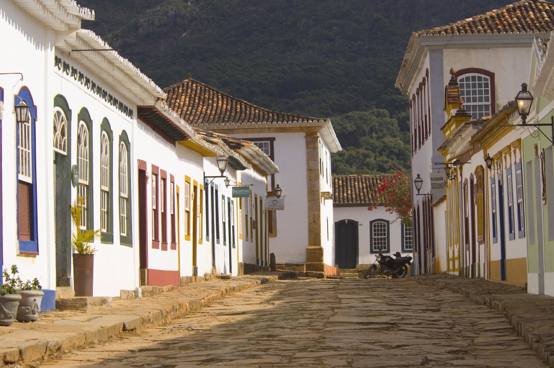 Colonial street in Tiradentes with white buildings, colorful doors and trim, forested mountains in background.