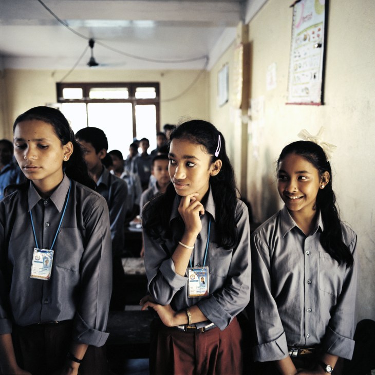Three female students in grey uniforms with ID badges stand in a school classroom.