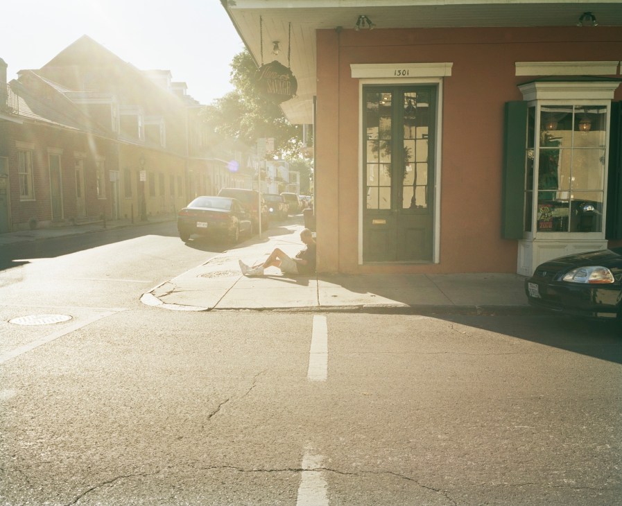 Sunlit New Orleans French Quarter street with colonial buildings, a parked car, and a person sitting on the pavement.