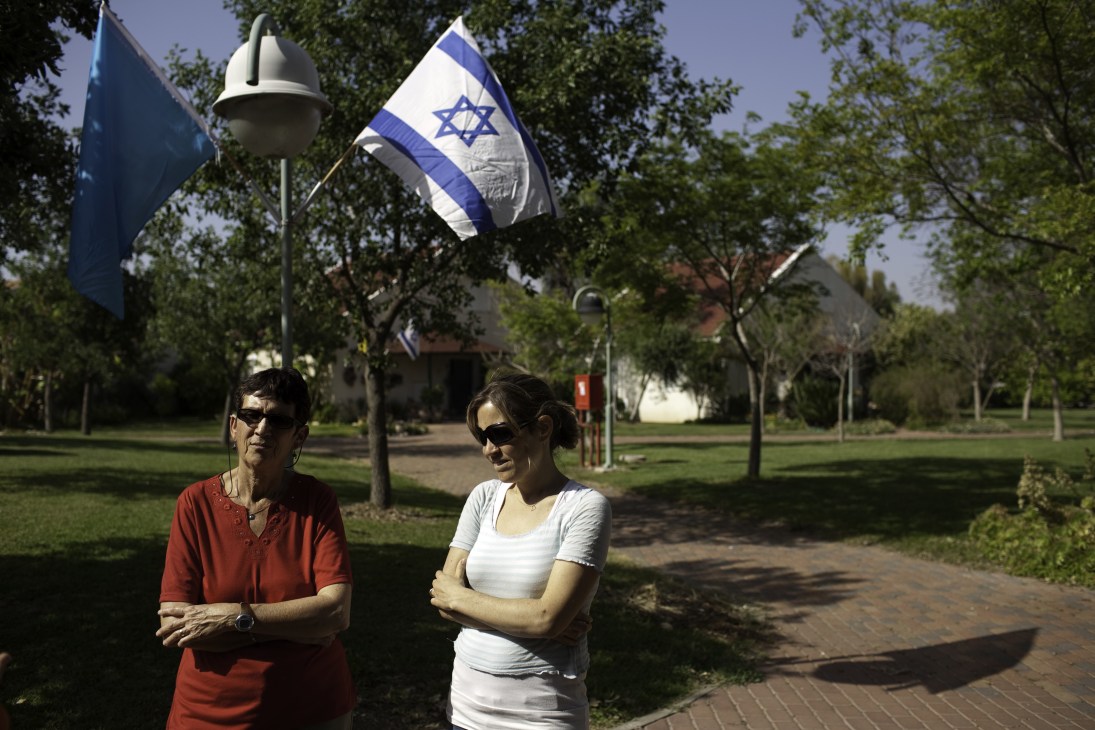 Two women stand on a tree-lined path beneath an Israeli flag at Maabarot kibbutz.