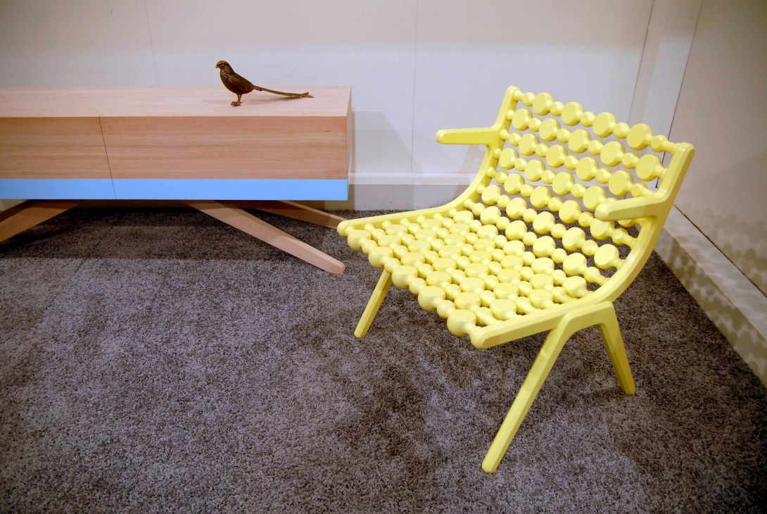 Bright yellow chair with knobbed seat design displayed alongside wooden furniture at Salone.
