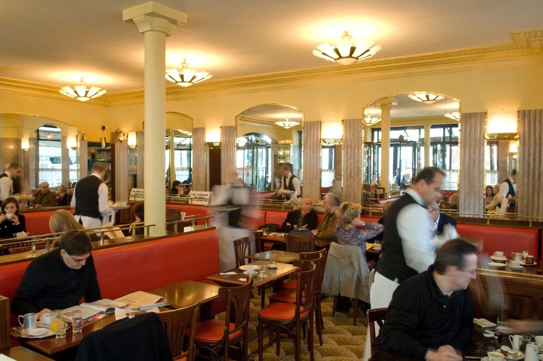 Art Deco interior of Café de Flore with cream-colored walls, supporting columns, brass fixtures, red banquettes, and patrons seated at tables.