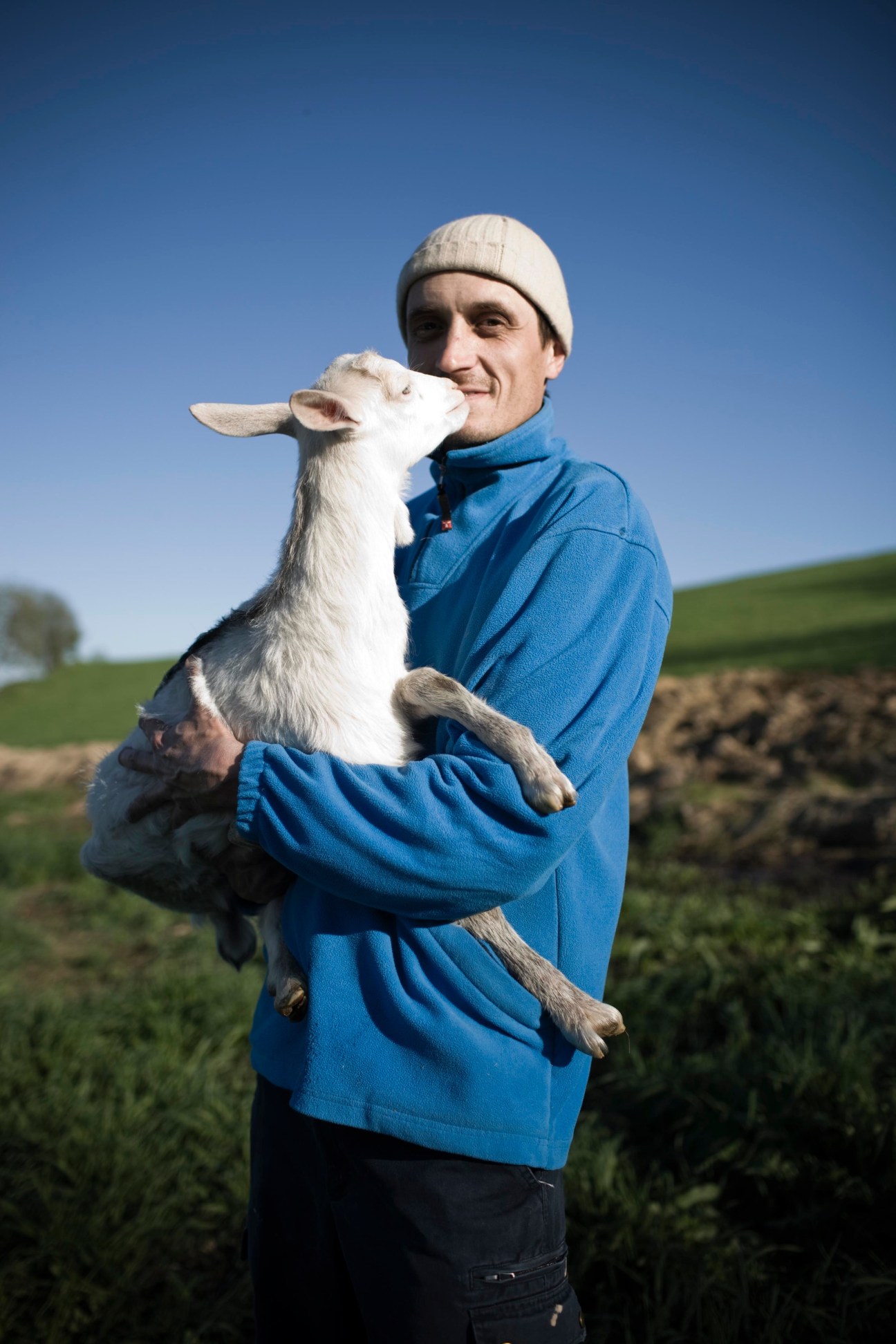 Farmer Giovanni Solerio holds a white young goat in his arms in a grassy field.