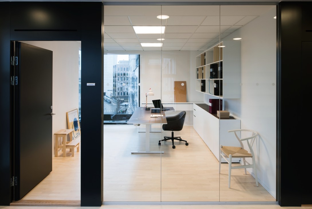 Modern individual office with glass walls, white desk, black chair, wooden shelving, and city views.