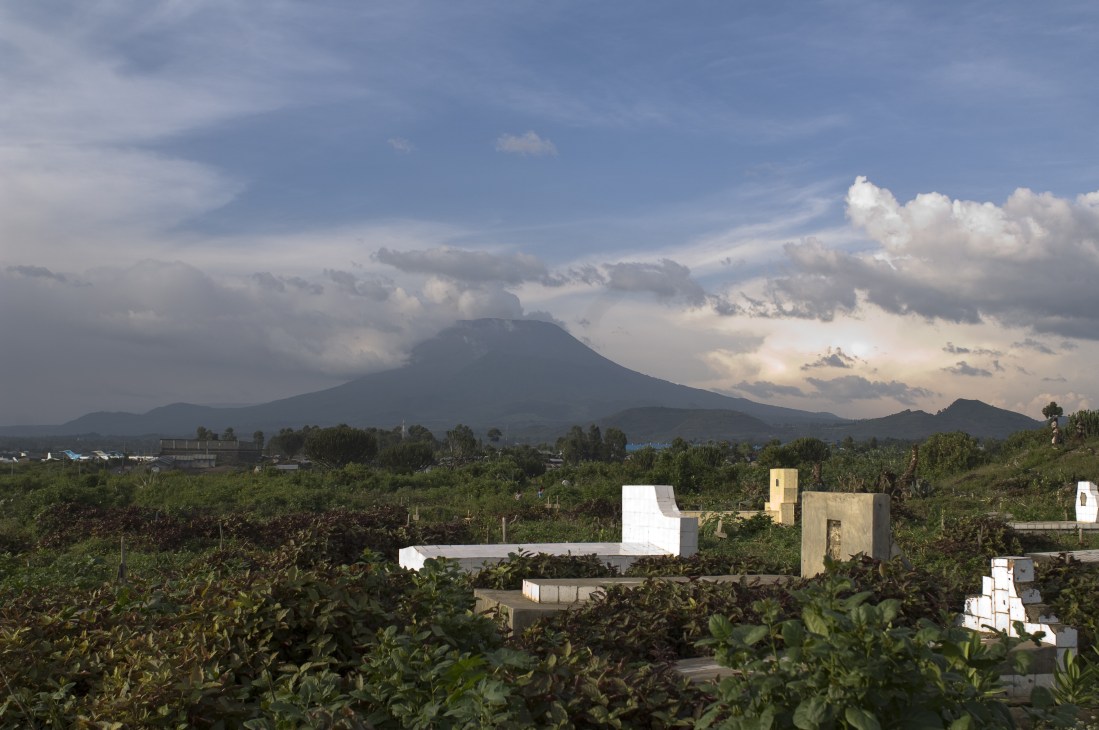 Nyiragongo volcano rises above a graveyard and green vegetation under cloudy skies in Goma.
