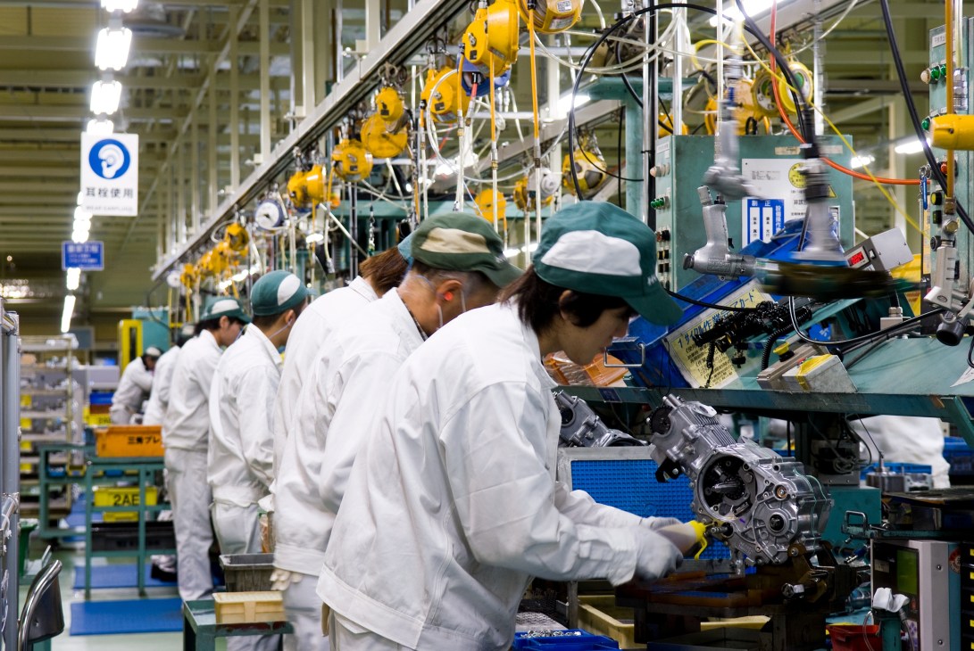 Workers in white coats and green caps assemble motorcycle engines on a factory production line with yellow overhead hoists.