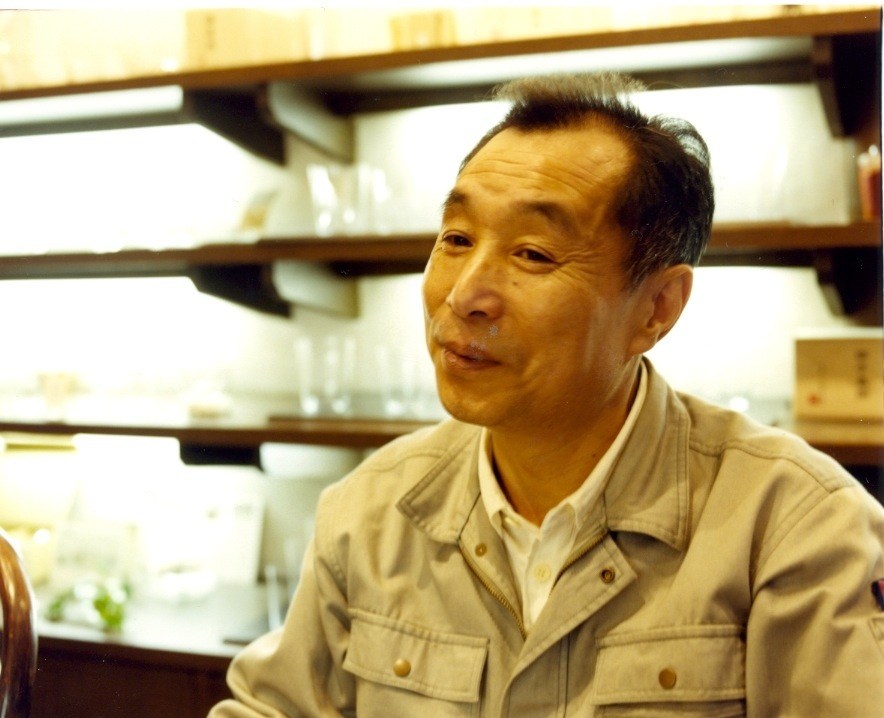 Kunio Muramatsu, president of Shotoku glassware company, seated indoors with shelving visible behind him.