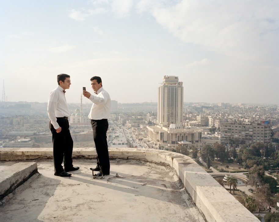 Two men in business attire stand on a rooftop terrace overlooking Aleppo's cityscape with a modern building visible in the distance.