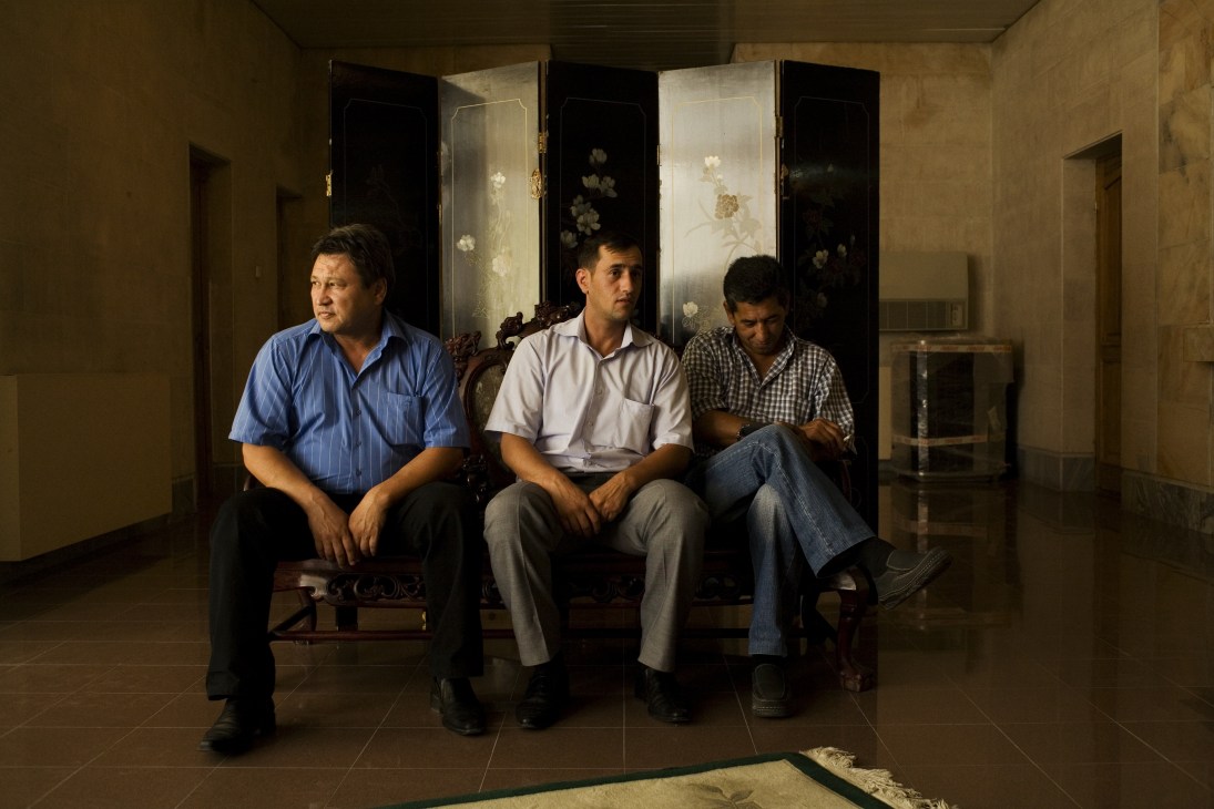 Three men sit on a bench in a hotel lobby with decorative panels behind them.