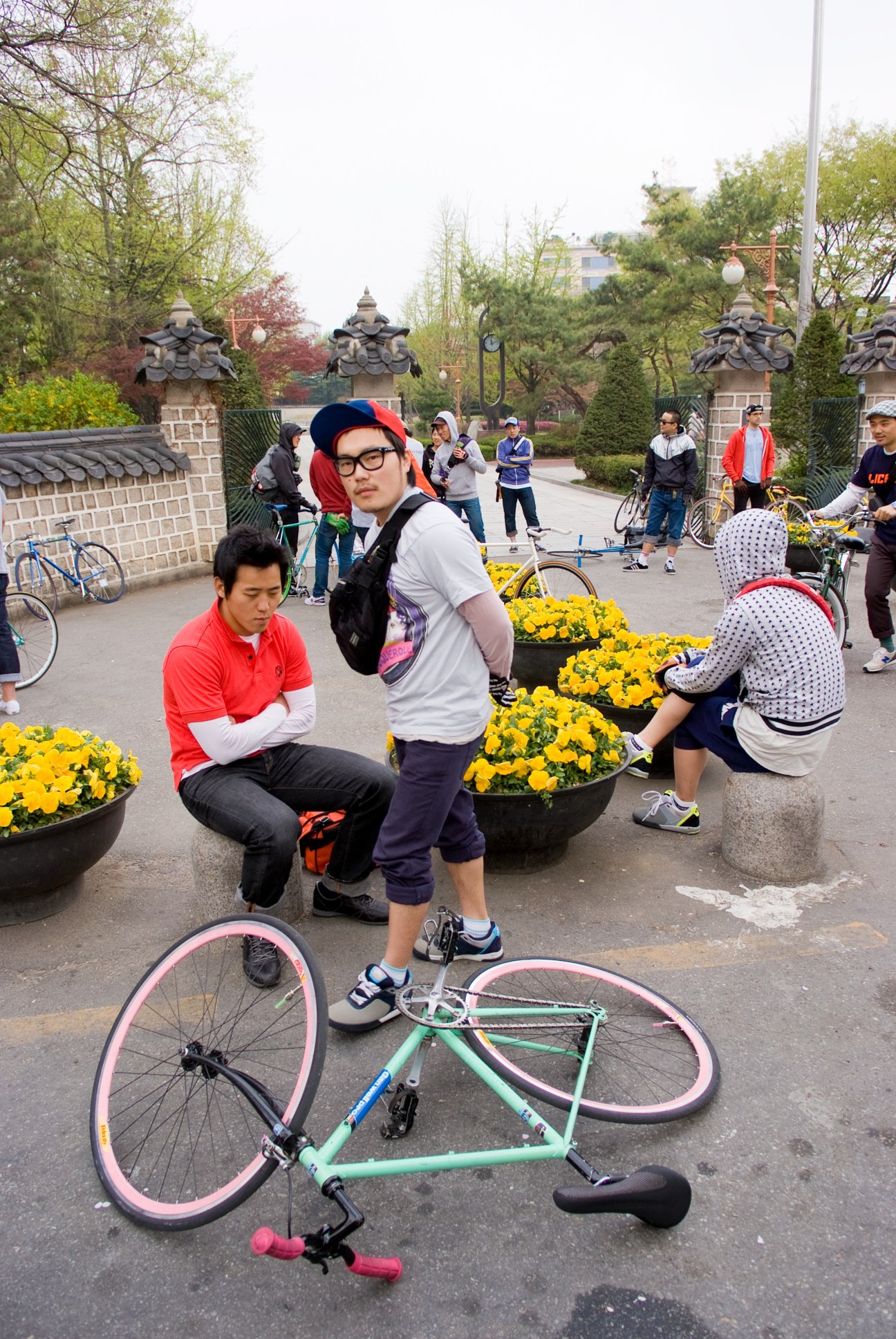 Cyclists gather at Dosan Park in Seoul with yellow flowers and traditional Korean architecture visible in the background.
