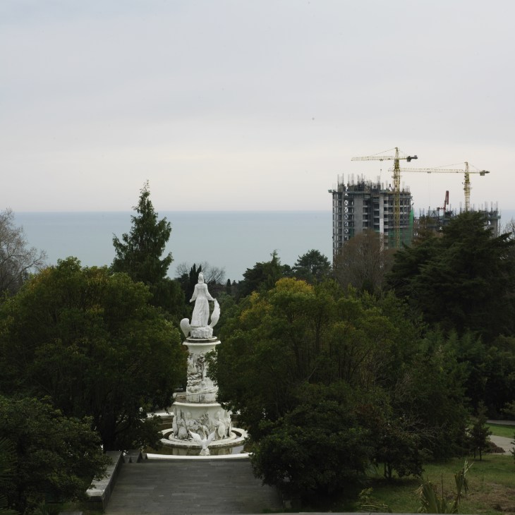White statue in Sochi's botanic gardens with Black Sea coast and construction cranes visible beyond.