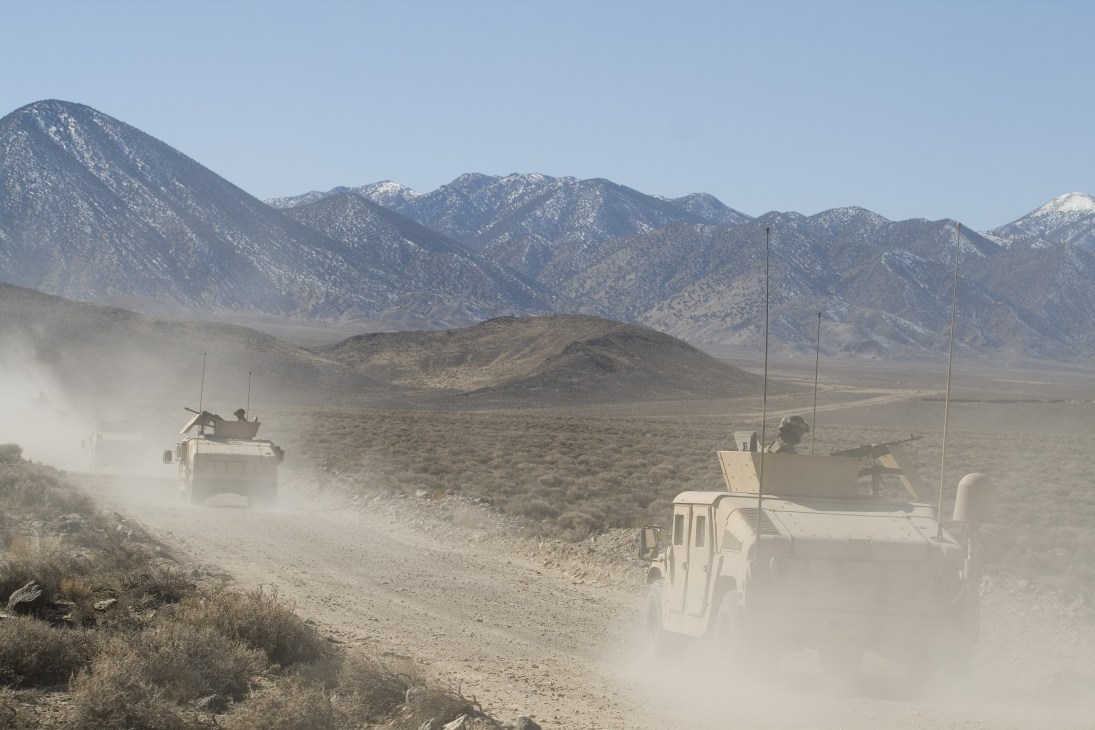 Armored Humvee convoy travels down a dusty desert road toward snow-capped mountains in Nevada.