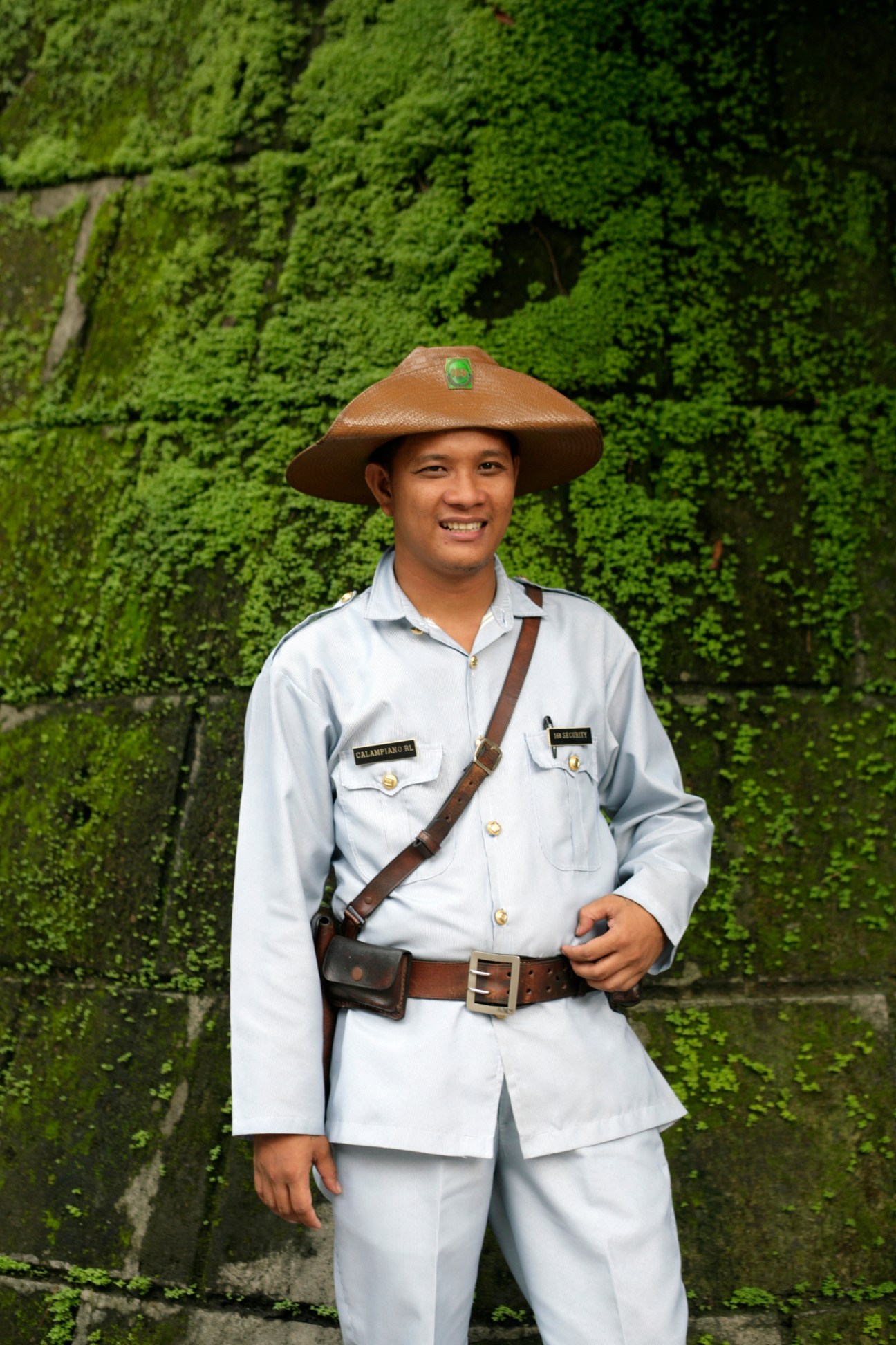 A uniformed guard in white stands smiling against ivy-covered stone wall at Intramuros, Manila's historic walled district.
