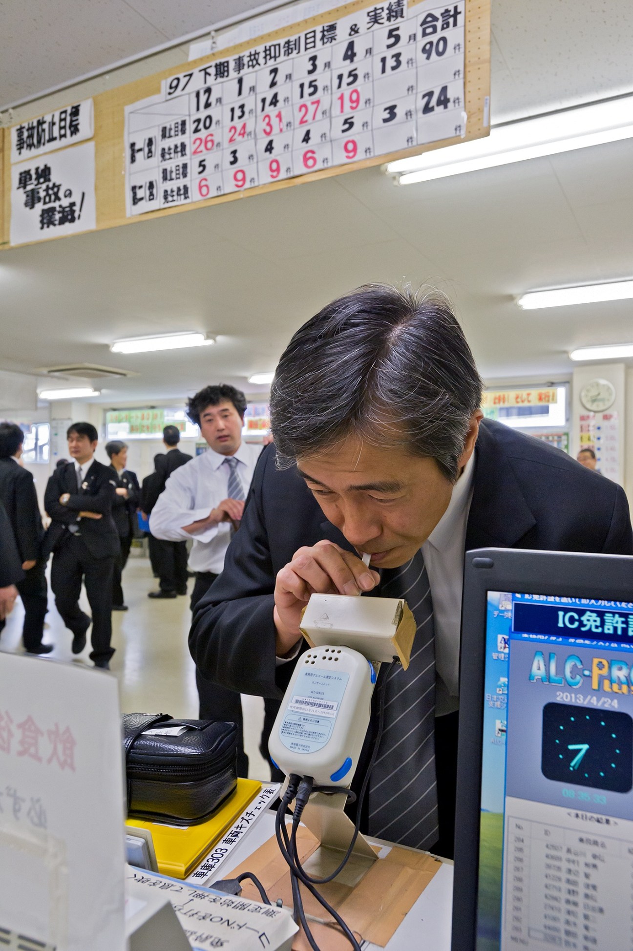 A taxi driver in dark suit takes a breathalyser test at Nihon Kotsu depot with schedule board and other staff in background.