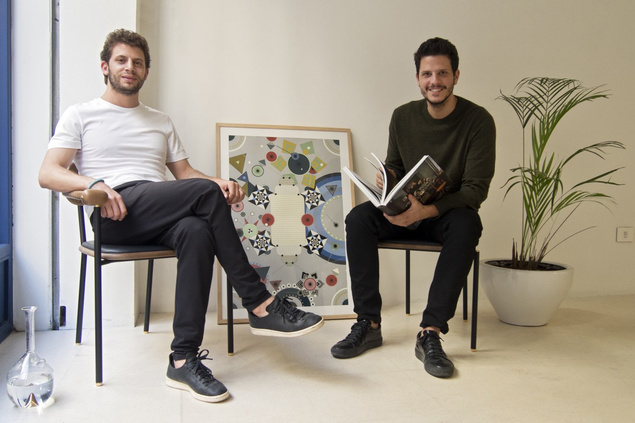 Designers Nicolas Moussallem and David Raffoul sitting in their studio with framed artwork and a potted plant.