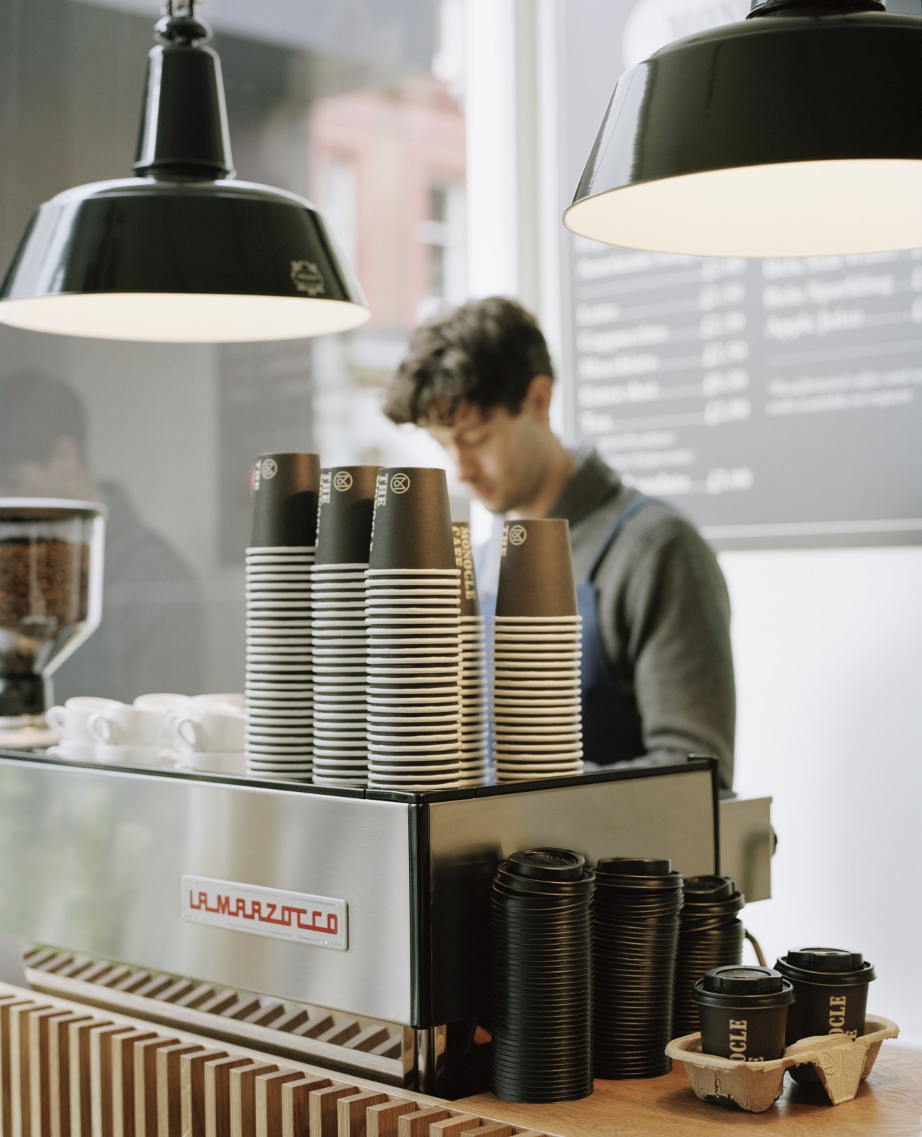 Barista at La Marzocco coffee machine with stacked paper cups and black pendant lights overhead.