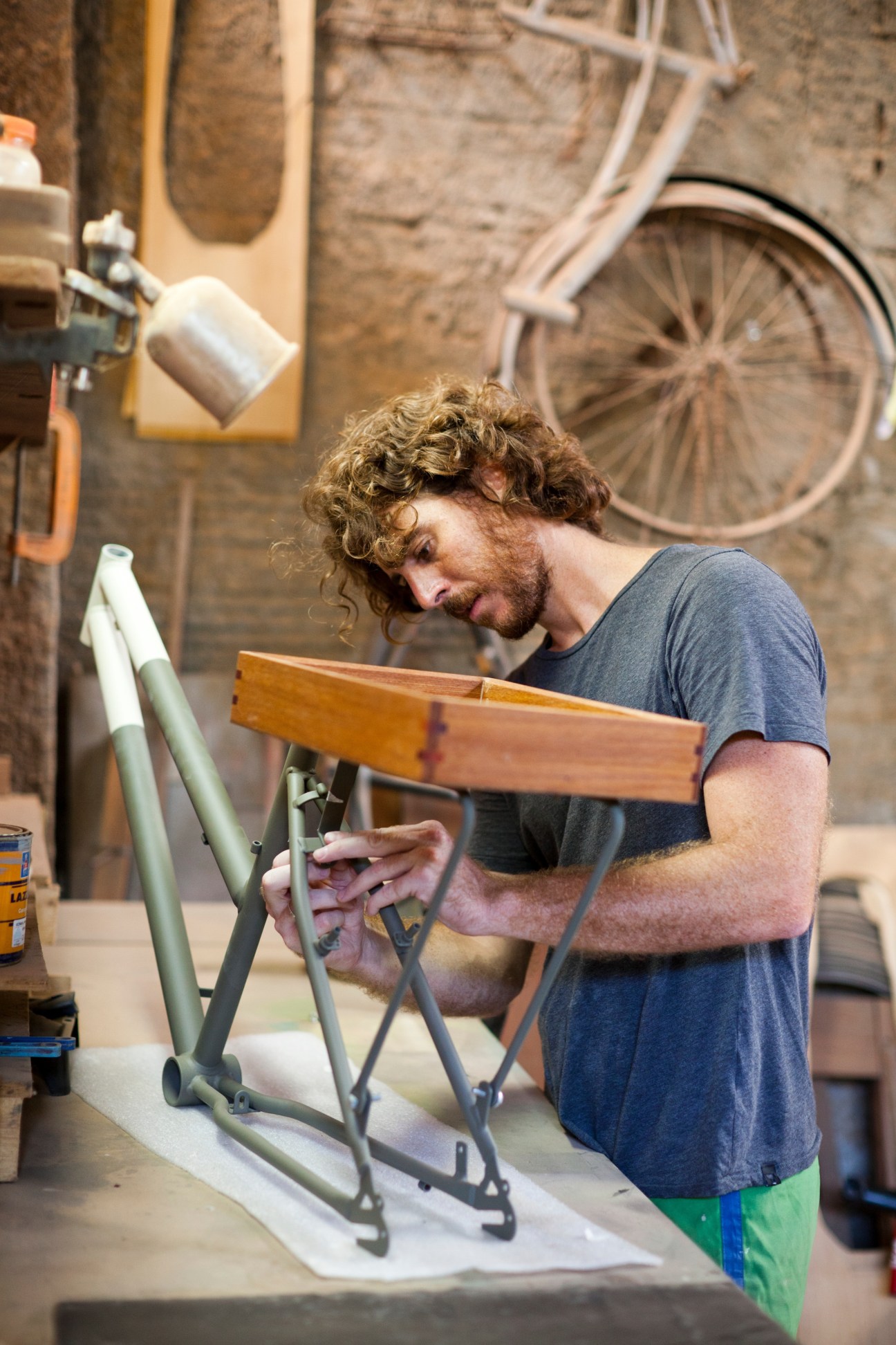 Man working on bicycle frame in workshop with finished bike mounted on wall behind him.