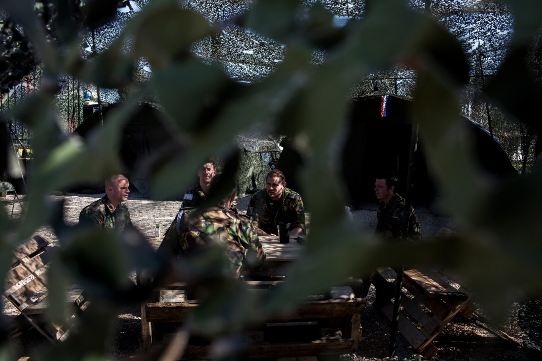 Dutch soldiers in camouflage uniforms sit at wooden tables under shade in a military camp.