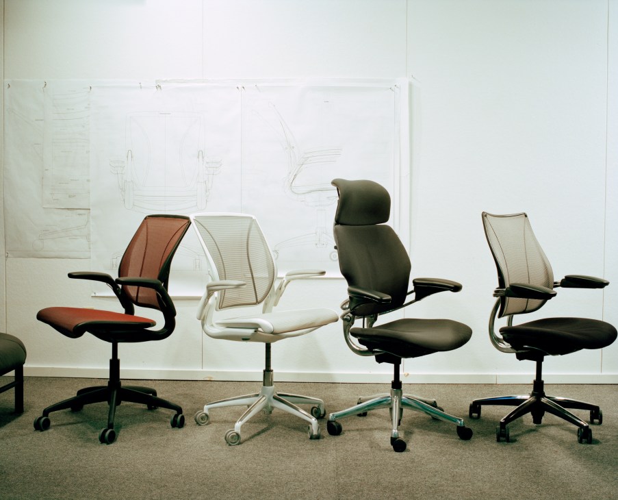 Four ergonomic office chairs in different colors displayed against a wall with design sketches.