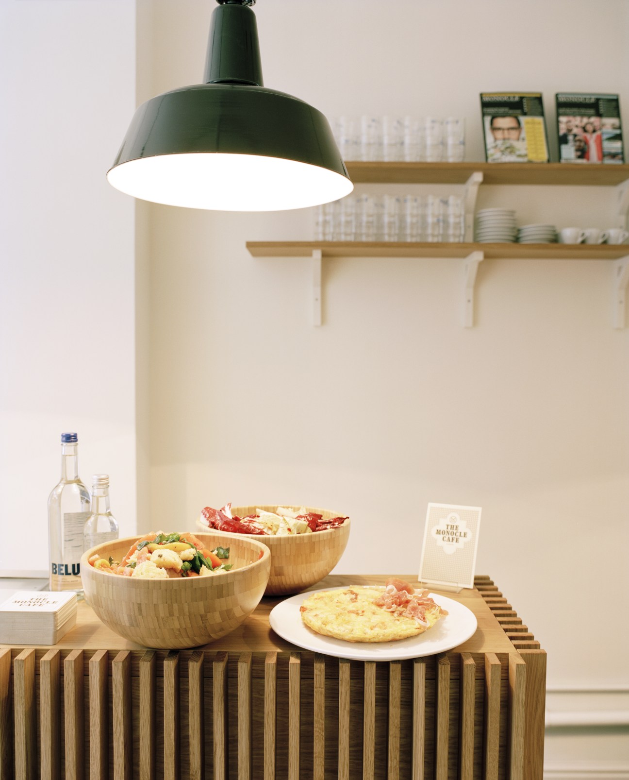 Wooden bowls with salad and frittata on a striped table under a green pendant lamp with shelving behind.