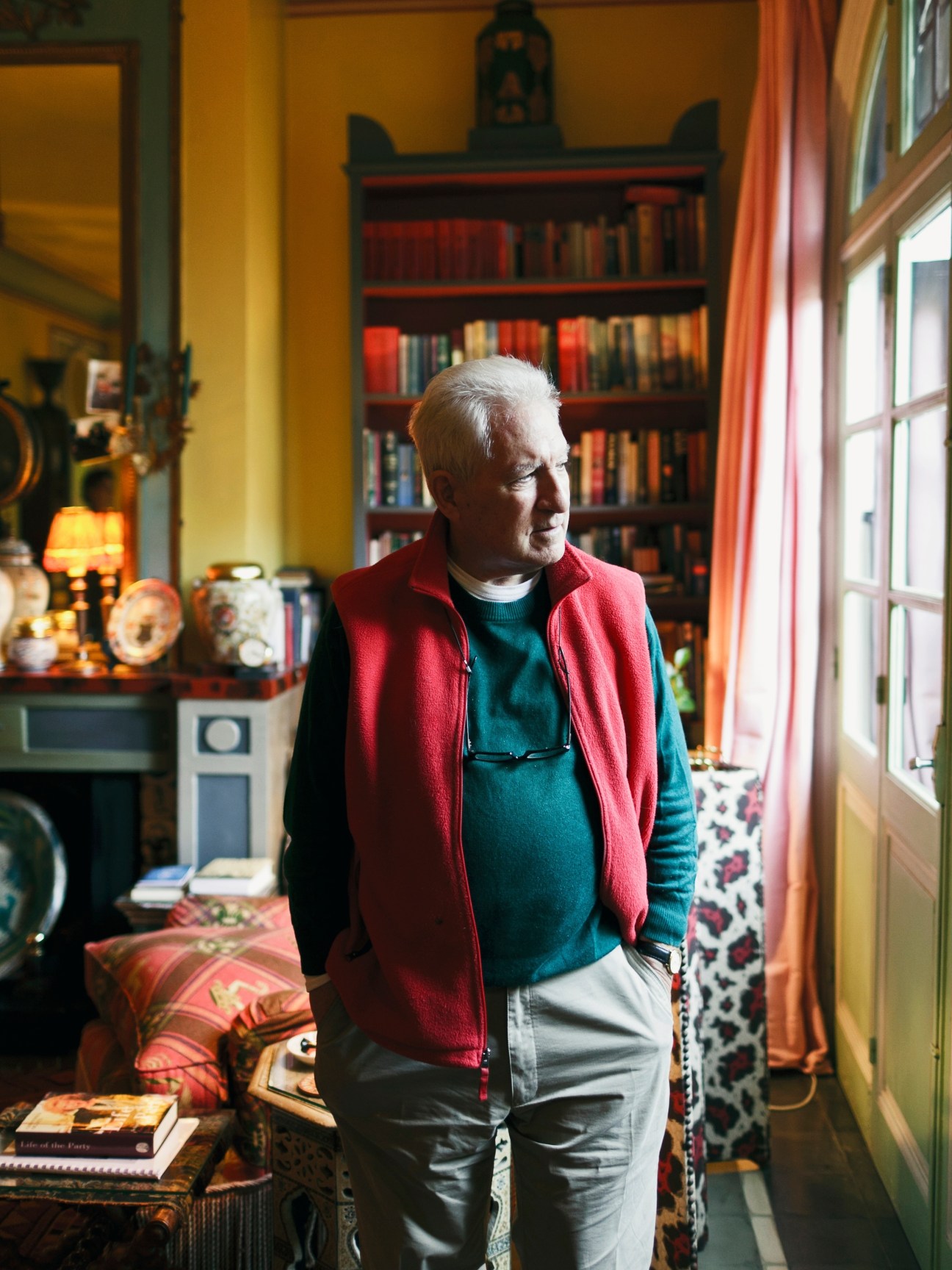 Older man with white hair wearing a red vest and teal sweater standing in a colorful interior with bookshelves and decorative objects.