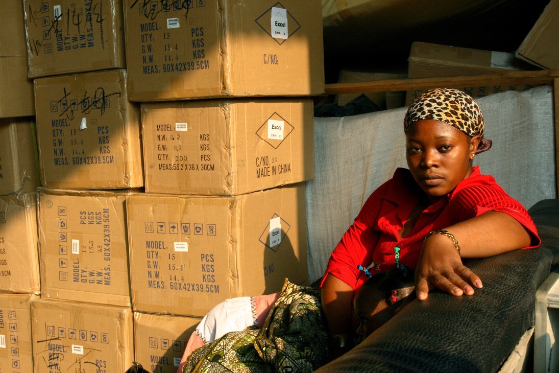African woman in red shirt and patterned headwrap sitting among cardboard shipping boxes marked 'Made in China'.