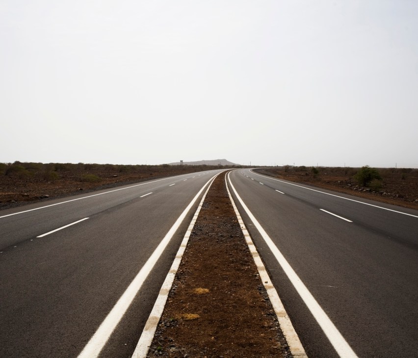 A straight new asphalt road with white line markings stretching across barren volcanic landscape under overcast sky.
