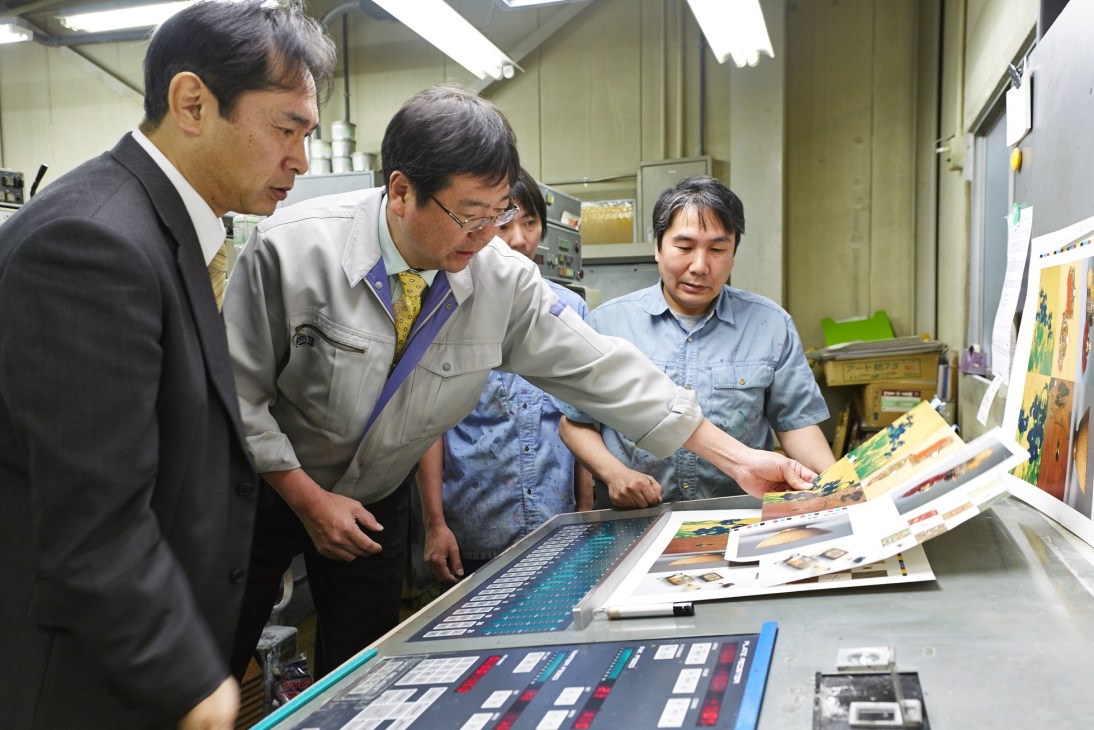 Men examining postcard proofs at a color correction station in a printing factory.