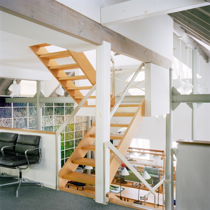 Interior view of a multi-level studio space with a wooden staircase, white walls, large windows overlooking greenery, and a black leather chair visible in the lower level.
