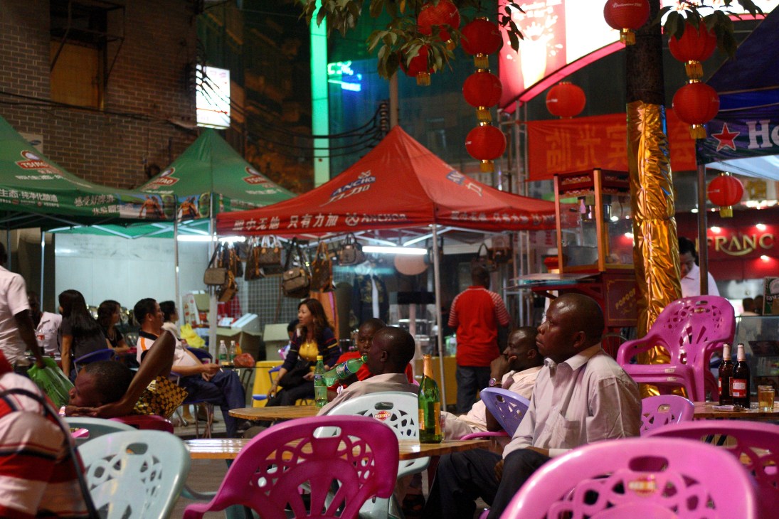 African patrons at an outdoor bar with red tent canopy, colorful plastic chairs, and Chinese lanterns in Guangzhou.