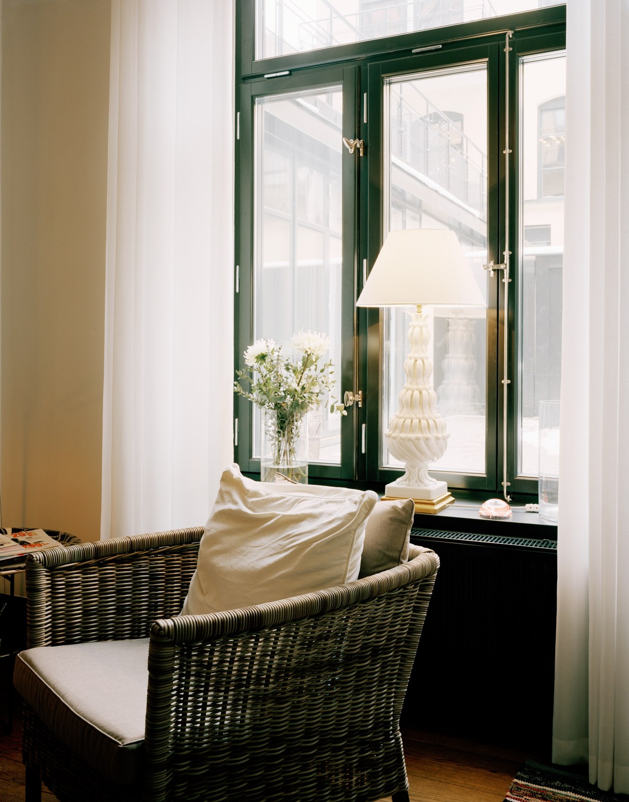 Wicker armchairs with cushions by a green-framed window with white lamp and flowers.