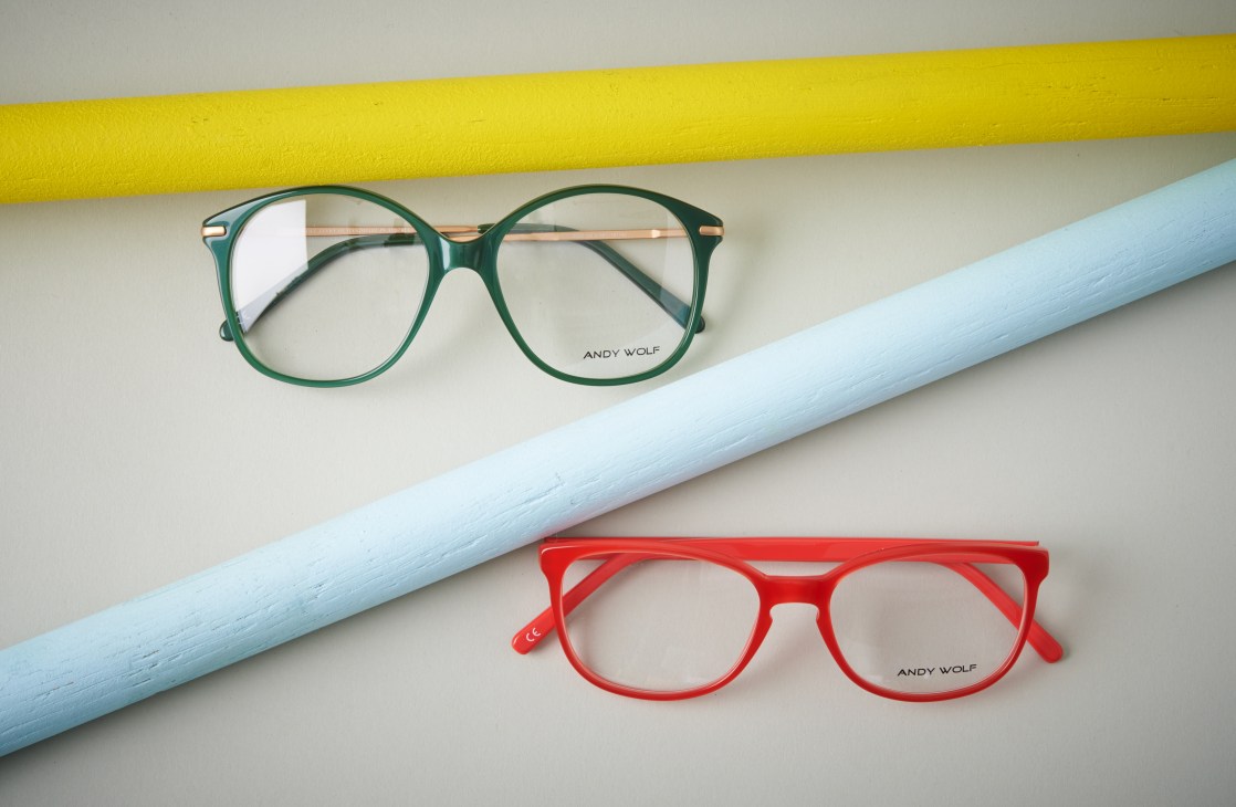 Green and red Andy Wolf eyeglasses displayed diagonally on a white surface with colored geometric shapes.