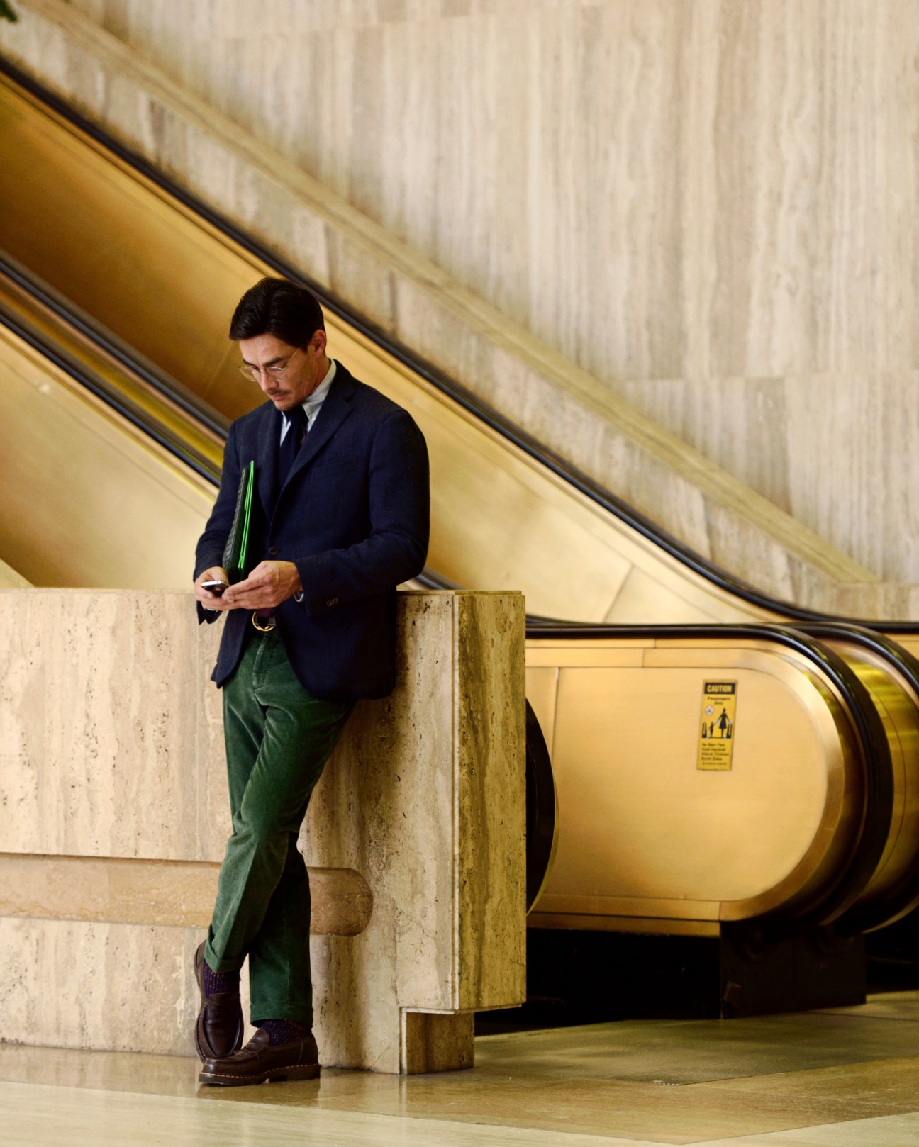 Man in navy Boglioli jacket and Incotex trousers standing in modern subway station holding green document case.