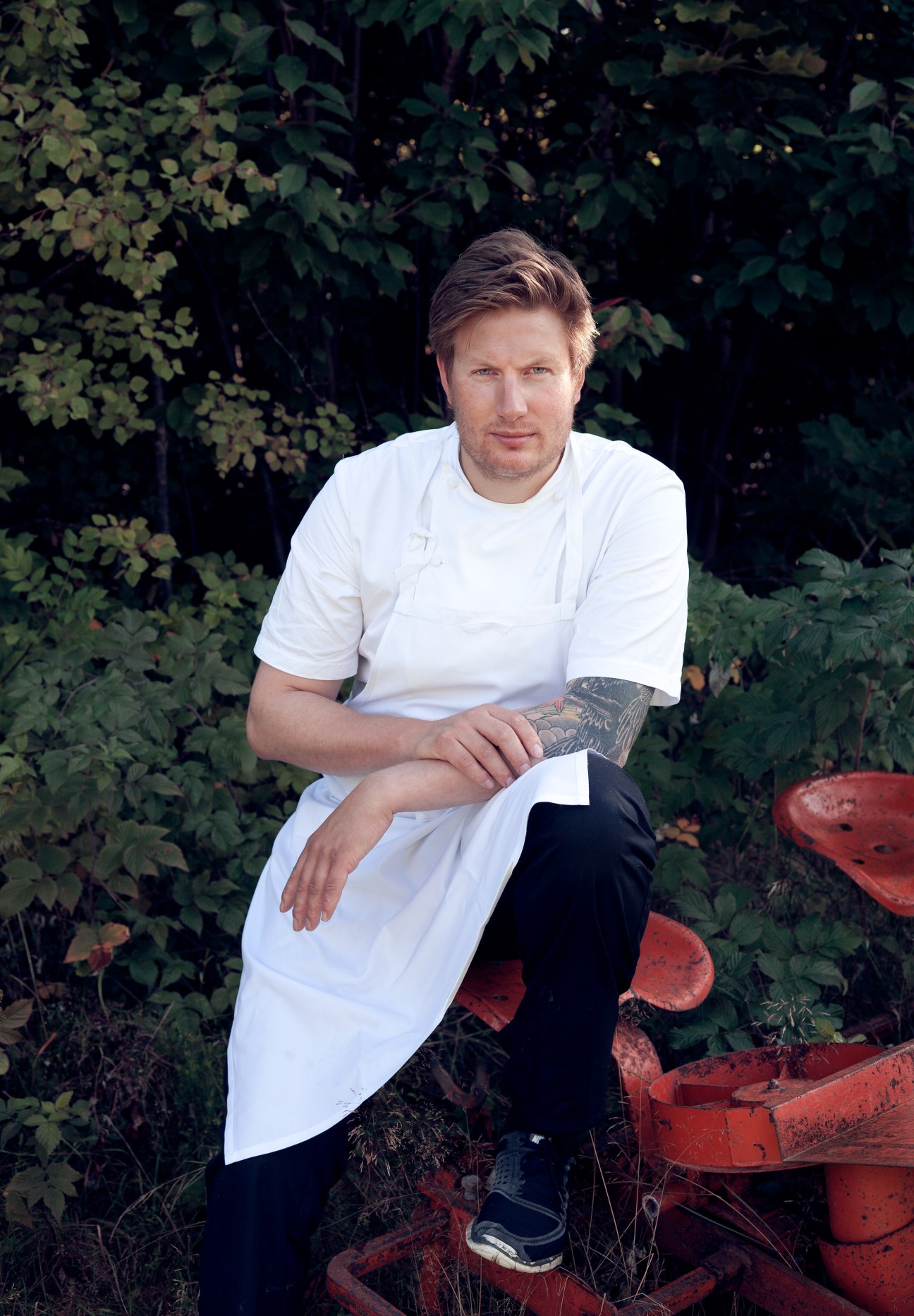 Chef Esben Holmboe Bang seated outdoors against foliage in a white chef's jacket.